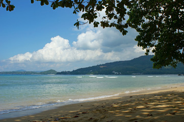 Coast of the Andaman sea. Sandy beach in the shade of a tree. Light waves roll ashore. Mountains in the background. A feeling of warmth and calm. Landscape from under a tree branch