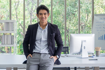 Portrait of an Asian businessman wearing a black suit, looking at the camera and smiling at the desk in the office.Garden background covered with green trees and copy space.
