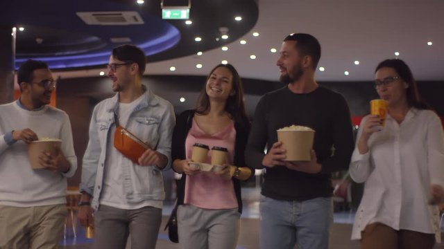 Tracking shot of happy friends eating popcorn and chatting while walking to watch movie at cinema