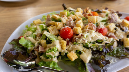 Mixed vegetables salad on white plate with caesar salad sauce, tomatos, red and green oak, crispy bread topping, food in dish on wooden table