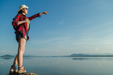 Asian young woman in a plaid shirt wearing a hat with a backpack by the lake on a summer holiday.