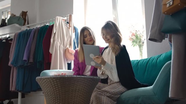 Optimistic young women in trendy outfits smiling and using tablet to select garments online while sitting on sofa in clothes store.