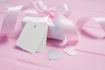 White gift box with a pink satin bow and ribbon on pink wooden background. Mock up with paper kraft tag is included for your text. Can use for Valentines Day, mothers day, birthday