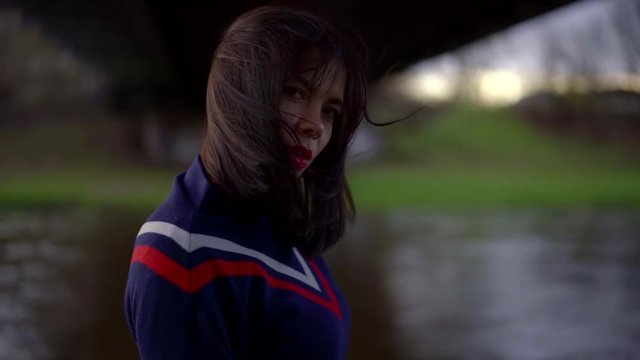 portrait of a brunette with bright lipstick on her lips and in a blue jumper on a cloudy day. she stands under the bridge by the river, her hair blowing in the wind