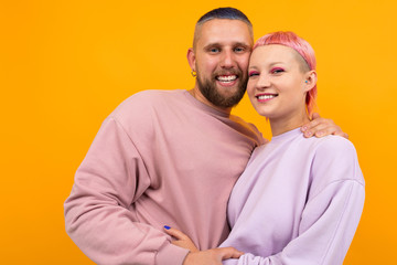 Unusual pretty woman with short pink hair and tattoo hugs with her boyfriend isolated on orange background