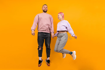 Unusual pretty woman with short pink hair and tattoo jumps with her boyfriend isolated on orange background.