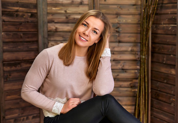 Loft style concept. Portrait of a pretty blonde girl in a pink sweater on a wooden background in the home interior. Cute looks and smiles at the camera, sitting on a chair straight