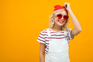 Beautiful young woman with short blonde curly hair and bright makeup in white overalls. red sunglasses and red hat gesticulated and smiles, portrait isolated on orange background