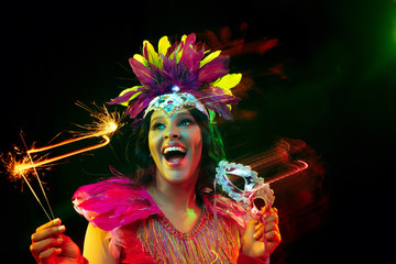 Beautiful young woman in carnival mask and stylish masquerade costume with feathers and sparklers in colorful lights on black background. Christmas, New Year, celebration. Festive time, dance, party. © master1305