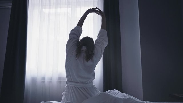 Woman wakes up, gets up on bed and stretches in morning sun light. Young woman greets new day