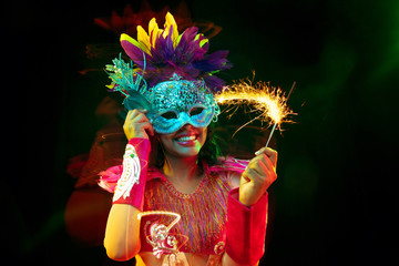 Beautiful young woman in carnival mask and stylish masquerade costume with feathers and sparklers in colorful lights on black background. Christmas, New Year, celebration. Festive time, dance, party. © master1305