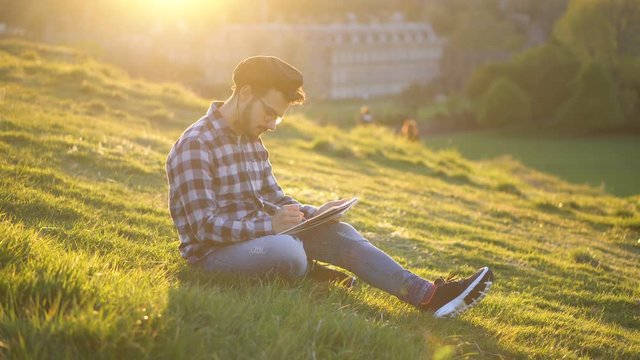 Talented Artist Drawing While Looking at Model or Subject Sitting on Meadow in Nature Environment With Sun Setting Flaring in Background
