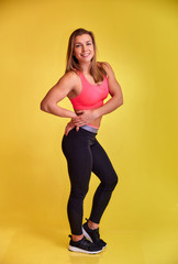 Vertical portrait of a pretty fitness blonde girl with a beautiful smile, excellent teeth and a bodybuilder figure in a tracksuit on a yellow background. Cute looks at the camera, standing straight.