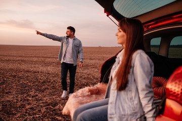 Young Happy Couple Dressed Alike in White Shirt and Jeans Sitting at Their New Car Trunk, Beautiful Sunset on the Field, Vacation and Travel Concept