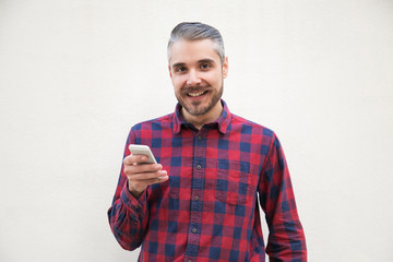 Content man holding smartphone and smiling at camera. Cheerful bearded man in checkered shirt using mobile phone and looking at camera on grey background. Technology concept