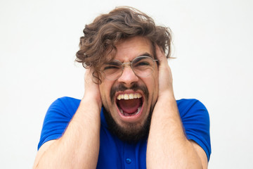 Nervous desperate guy covering ears with hands and shouting loud. Handsome bearded young man in blue casual t-shirt posing isolated over white background. Stress concept