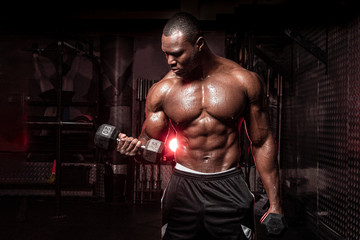 Muscular African American shirtless male bodybuilding athlete does dumbbell curls  in a dark grungy gym with dramatic lighting flare 