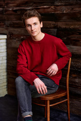 Loft style concept. Portrait of a handsome young man. Short-haired Caucasian in a burgundy sweater on a vintage wood background. Cute looks at the camera sitting on a chair.