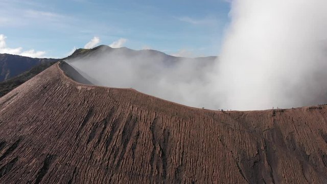 Aerial shot of smoking Mount Bromo, shot of tourists walking on pathway on crater edge, slowly flying backwards shot, horizon view. Java, Indonesia