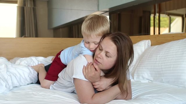 Portrait of a young woman with her son at home on the bed, the child sits on the back of her mother and hugs her. Happy mom with baby having fun in the morning on the bed. Slow motion.