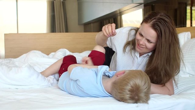 Portrait of a young woman with her son at home on the bed, a mother tickles her son and they laugh. Happy mom with baby having fun in the morning on the bed. Slow motion.
