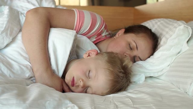 Close-up of mom gently hugging her sleeping son in the morning in bed. Mother and child sleep together in a white bed. Slow motion.