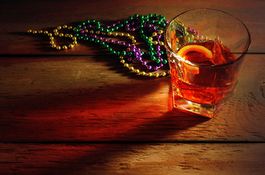 Close-up View Of Sazerac Cocktail With A Lemon Peel In Glass On Wooden Table With Side Lighting And Warm Tones. Mardi Gras Beads In Green, Gold And Purple Nearby.