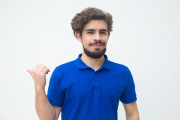 Fototapeta premium Positive smiling guy pointing thumb away at copy space. Handsome bearded young man in blue casual t-shirt posing isolated over white background. Advertising or sharing news concept