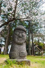 single Dol Harubang (Hareubang) statue in green grassland and cherry trees in background at Jeju Island, South Korea, Asia 