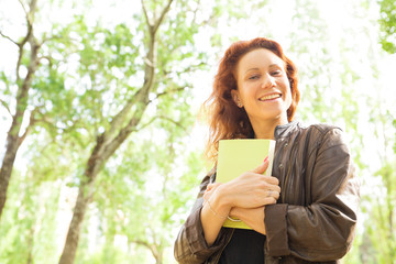 Fototapeta premium Happy joyful beautiful woman walking in park, carrying book. Red curly haired young woman in casual posing outdoors. Book reader concept