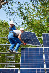 Two workers mounting heavy solar photo voltaic panel on tall steel platform standing on ladder on green tree and blue sky background. Exterior solar system installation, dangerous job concept.