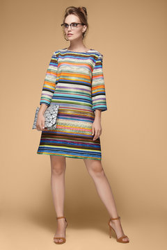 A charming European blonde lady in a colorful striped gown is posing on a beige background. The girl with elegant glasses and hair bun is holding a silver geometric clutch in her hand.