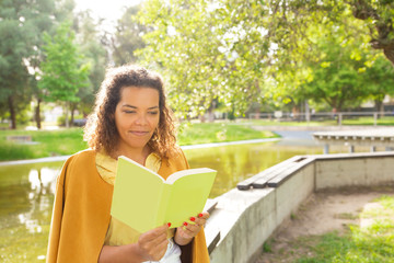 Fototapeta premium Pensive woman enjoying reading in park, sitting on parapet near river. Green grass background. Curly haired black woman in casual posing outdoors. Reading book, leisure concept