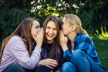 Fototapeta premium Three young attractive woman sharing secrets sitting on green grass in the park. Cheerful girlfriends gossip and whisper outdoor.