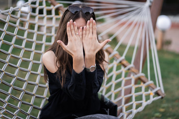 Happy woman in dark shirt, jeans and sunglasses showing a ring and relaxing in hammock in forest at...