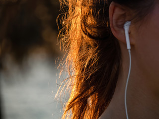 A girl with beautiful sunlight in hairs is listening music with earphones. No face.