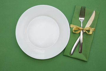 empty white plate with fork and knife and green napkin on a green background. Festive table setting, serving, catering. Copy space.
