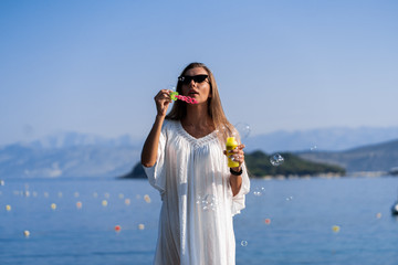 Young beautiful woman in white dress and sunglasses blowing soap bubbles on pier with seaview background. The concept of joy, ease and freedom during the vacation. The girl is enjoying the rest.