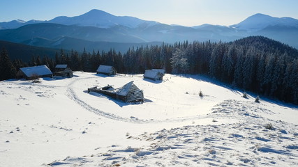 Panoramic view of the snow capped mountains and pine forest. Cold sunny day in winter mountains. Small village with shepherd houses. Carpathians, Ukraine.