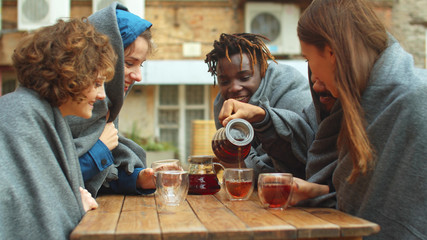 Friends in blankets sit outside the cafe and pour tea in cups