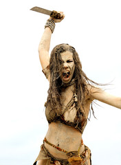 A young woman is  dressed as a neanderthal warrior.  She is covered with mud, filth and dirt and is...