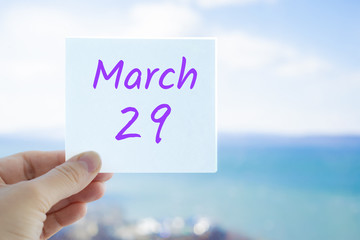 March 29th. Hand holding sticker with text March 29 on the blurred background of the sea and sky. Copy space for text. Spring month in calendar concept