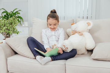 Cute little girl sitting on the sofa with teddy-bear near her, reading fairy-tale, pointing her finger at something in the book, smiling.