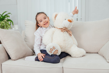 Smiling little kid is sitting on the sofa with white plush bear, looking at the camera, giving her thumb up.