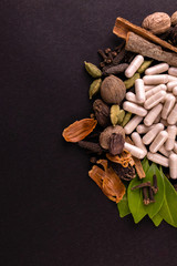 Herbal capsules with green leaves and assorted spices on black background in portrait mode. Herbal concept