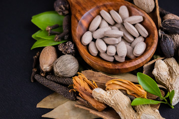 High angle front view of brown herbal tablets with green herbs and dry spices on black background. Alternative medicine concept