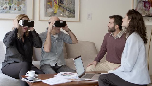 Two architects talking with clients in office. Smiling couple sitting on sofa with virtual reality headsets and gesturing. Technology concept