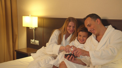 Parents and son sit on bed and look at photo on phone in hotel room