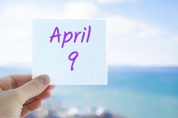 April 9th. Hand holding sticker with text April 9 on the blurred background of the sea and sky. Copy space for text. Spring month in calendar concept