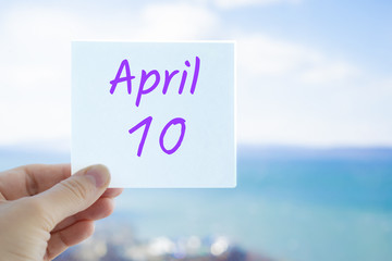 April 10th. Hand holding sticker with text April 10 on the blurred background of the sea and sky. Copy space for text. Spring month in calendar concept